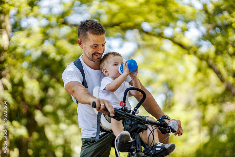 A boy drinking refreshment while sitting on a bike with his father.