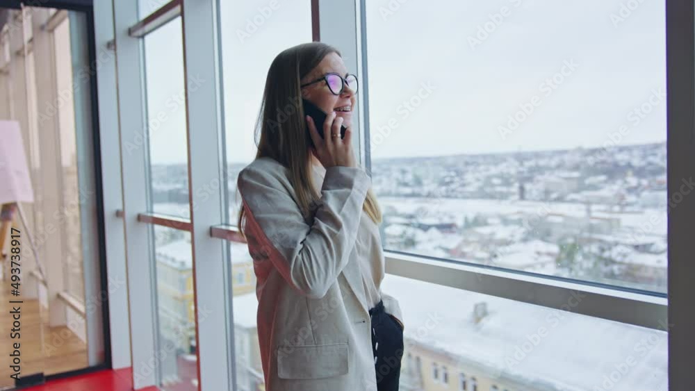 Attractive lady in glasses stands at the window speaking on the phone. Pleasant phone conversation in spacious office. Attractive lady in glasses stands at the window speaking on the phone. Pleasant phone conversation in spacious office.