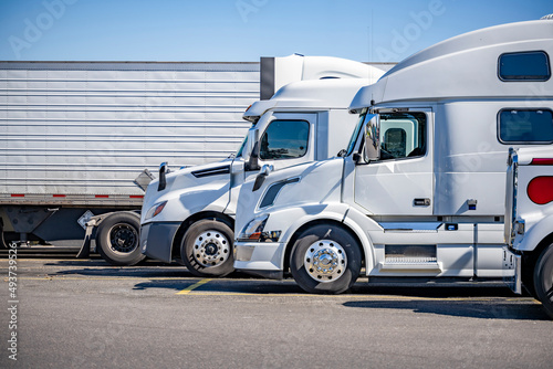 Profiles of different white big rigs semi trucks with semi trailers standing on the truck stop parking lot taking a break according to the schedule of cargo movement