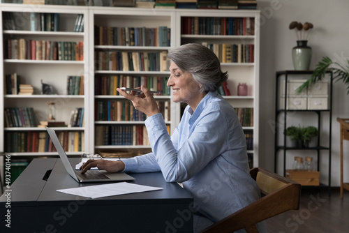 Older generation using modern tech, comfort communication, ai, workflow concept. Mature 60s business woman sit at workplace desk with laptop holds cellphone makes call talks to client on speakerphone
