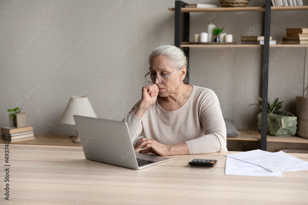 Aged woman sit at desk with calculator and papers, pay bills through e-bank application, looks at laptop screen feels concerned due bank debt or overdue. Older gen use tech, finances, troubles concept Aged woman sit at desk with calculator and papers, pay bills through e-bank application, looks at laptop screen feels concerned due bank debt or overdue. Older gen use tech, finances, troubles concept