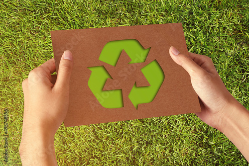 Hand holding a cut out paper with Symbolic of recycling and reuse shape on green grass field background.