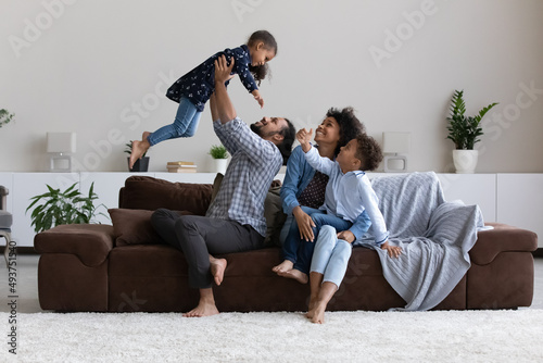 Cheerful excited young African parents having fun with little kids, enjoying leisure, activity on couch at home. Happy dad lifting joyful daughter girl up in air, mom hugging son. Family playtime