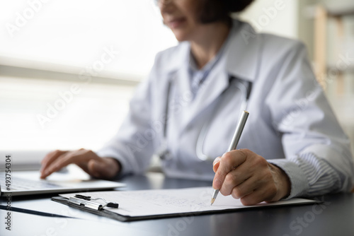 Hands of mature doctor woman filling paper medical records. Practitioner in white coat doing paperwork at workplace with laptop, writing notes, preparing documents, reports, prescription. Close up photo