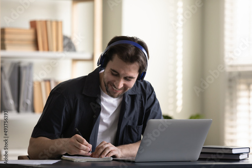 Cheerful student guy in headphones enjoying online studying at laptop, listening audio lecture, class, lesson, workshop, watching learning webinar, writing notes at computer, smiling, laughing