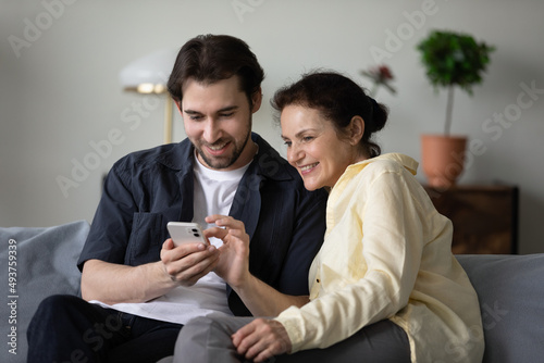 Happy positive grown son and mature mom using online virtual app, banking service on mobile phone, shopping on Internet together, making video call. Middle aged mother and grown child using gadget