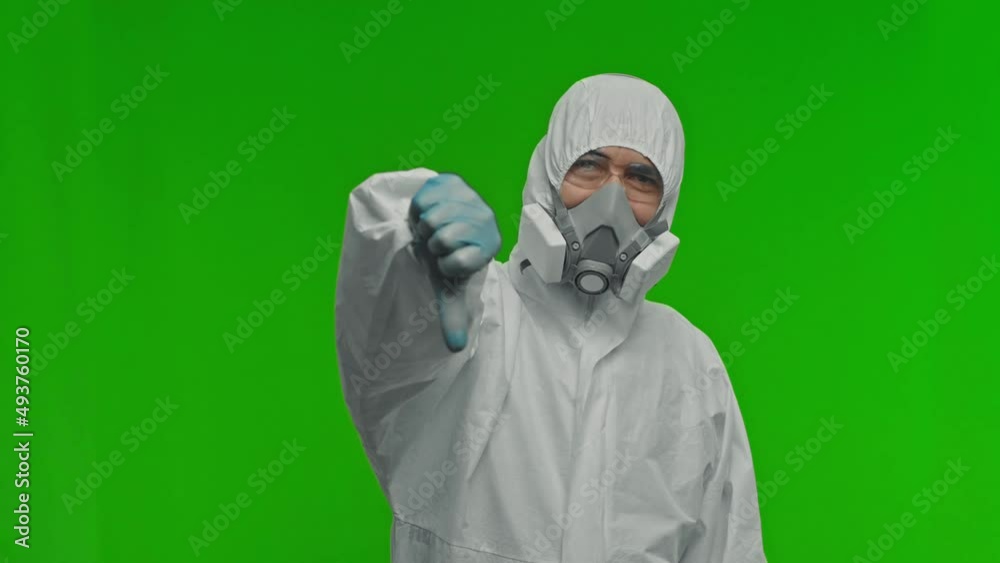 Serious man doctor wearing protective suit, goggles and respirator is showing thumbs down gesture. Concept of Coronavirus COVID-19 Pandemic. Studio shot in slow motion on isolated green background. Serious man doctor wearing protective suit, goggles and respirator is showing thumbs down gesture. Concept of Coronavirus COVID-19 Pandemic. Studio shot in slow motion on isolated green background.