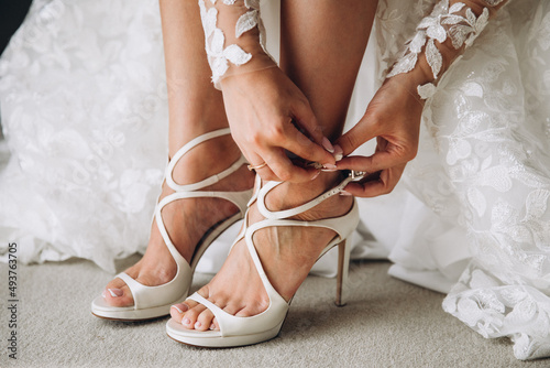a young bride in a white wedding dress sits and puts on white festive high-heeled shoes