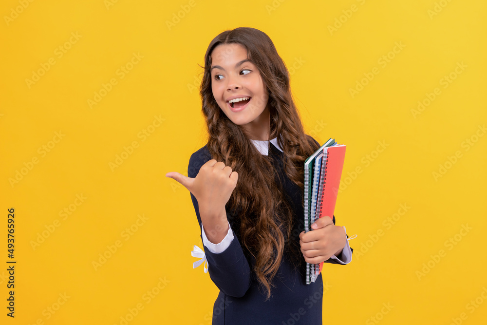 happy teen girl directing with thumb up hold school copybooks on yellow background, education. happy teen girl directing with thumb up hold school copybooks on yellow background, education.