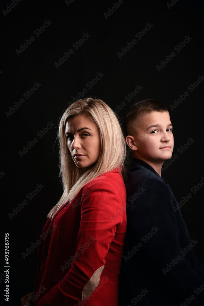 A young attractive mother stands back to back with her teenage son. Studio photo on a dark background
