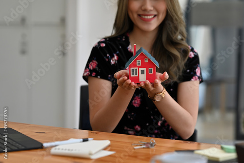 Cropped view young pretty Asian woman smiling, sitting in the office and holding a red house model, for housing, finance and real estate business.