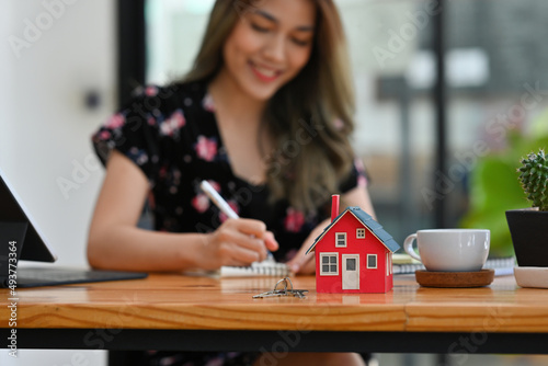 Cropped view young pretty Asian woman sitting in the office working, having a key and a house model in front, for real estate, business, finance and technology concept.