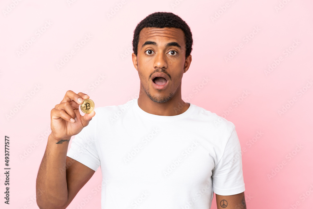 Young African American man holding a Bitcoin over isolated pink background with surprise facial expression Young African American man holding a Bitcoin over isolated pink background with surprise facial expression