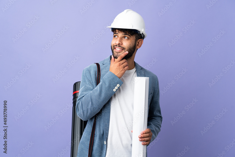 Young architect Moroccan man with helmet and holding blueprints over isolated background looking up while smiling Young architect Moroccan man with helmet and holding blueprints over isolated background looking up while smiling