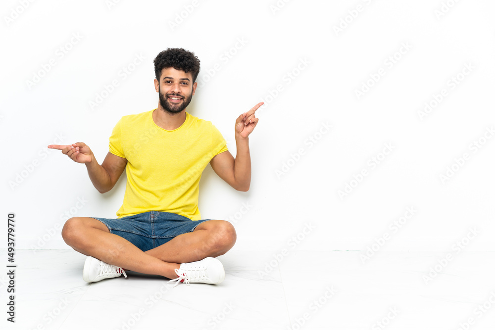Young Moroccan handsome man sitting on the floor over isolated background pointing finger to the laterals and happy Young Moroccan handsome man sitting on the floor over isolated background pointing finger to the laterals and happy