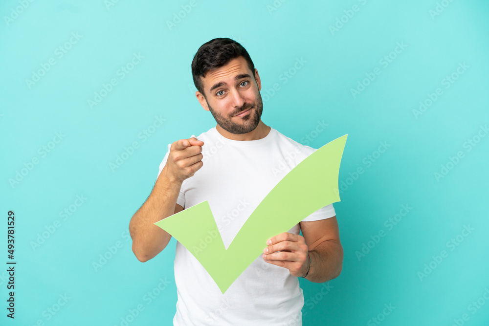 Young handsome caucasian man isolated on blue background holding a check icon and pointing to the front Young handsome caucasian man isolated on blue background holding a check icon and pointing to the front
