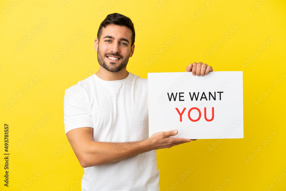 Young handsome caucasian man isolated on yellow background holding We Want You board with happy expression Young handsome caucasian man isolated on yellow background holding We Want You board with happy expression