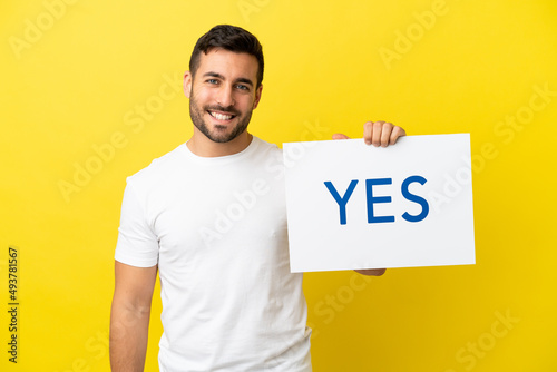 Young handsome caucasian man isolated on yellow background holding a placard with text YES with happy expression