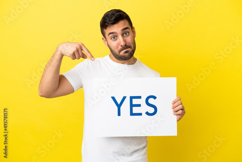 Young handsome caucasian man isolated on yellow background holding a placard with text YES and pointing it