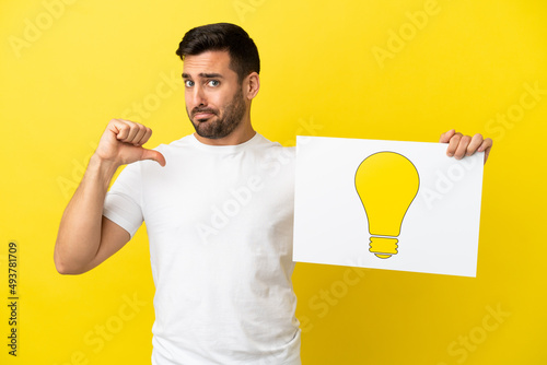 Young handsome caucasian man isolated on yellow background holding a placard with bulb icon with proud gesture
