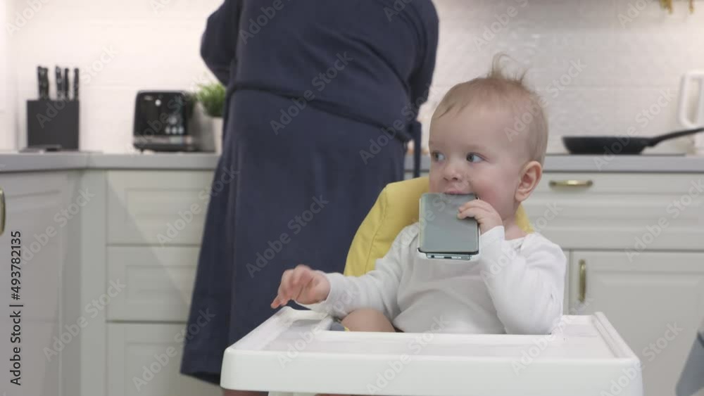 Father handing mobile device to child to entertain him, baby boy holding mobile phone sitting on high chair in the kitchen. High quality 4k footage Father handing mobile device to child to entertain him, baby boy holding mobile phone sitting on high chair in the kitchen. High quality 4k footage