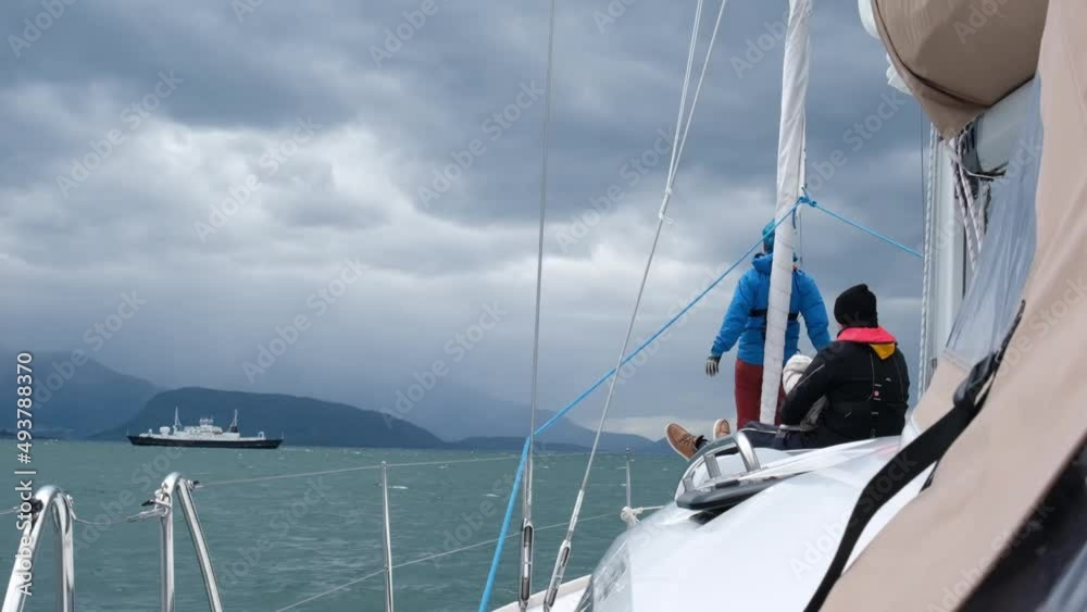 Two people on board a sailing yacht look out into the overcast sea and a passing ship. Mountains and overcast sky with clouds on the horizon. Two people on board a sailing yacht look out into the overcast sea and a passing ship. Mountains and overcast sky with clouds on the horizon.