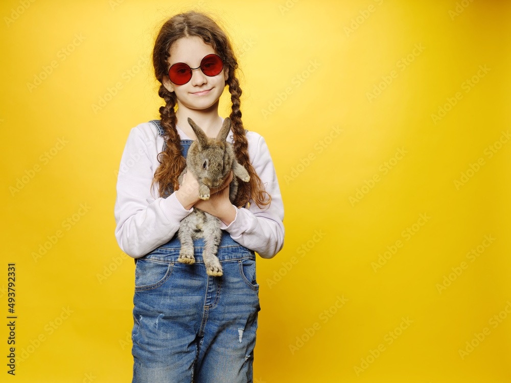 Front view of a little girl in a jeans and red sunglasses, holding a real grey rabbit, isolated yellow background. Horizontal view.
