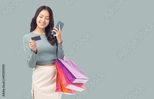 Young asian lady using mobile phone holding credit card carry shopping bags isolated over pastel green background and copy space Smiling young girl purchasing online through phone using credit card