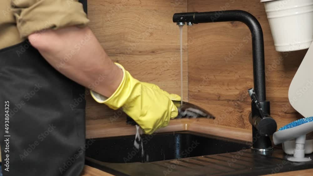Man in apron washes black plate with sponge and detergent under water running from faucet. Householder does chores standing in kitchen, closeup Man in apron washes black plate with sponge and detergent under water running from faucet. Householder does chores standing in kitchen, closeup