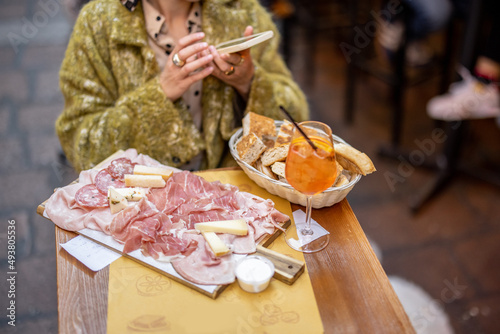 Woman photographing plate with Italian meat appetizer and Spritz Aperol drink while sitting at restaurant outdoors in Bologna city. Concept of Italian gastronomy and lifestyle