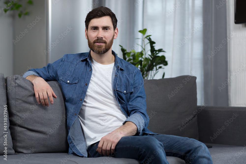 Portrait of a man with a beard at home, sitting on the couch and looking at the camera Portrait of a man with a beard at home, sitting on the couch and looking at the camera