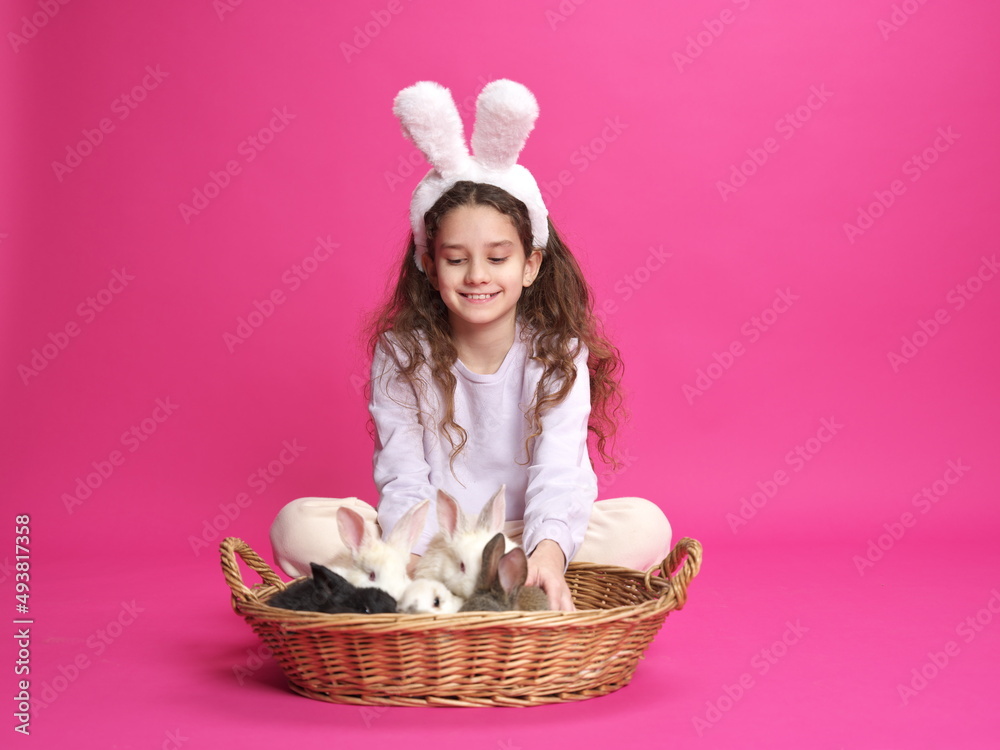 Full length image of a smiling little girl with white bunny ears, showing straw basket with little rabbits, isolated on a pink background.