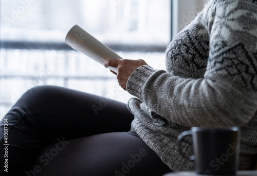 A woman holds a book in her hands and reads near the window.It's very bad weather outside