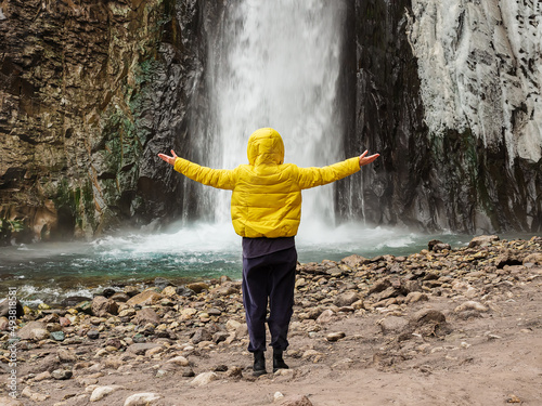 A woman in a yellow jacket stands on a rocky river bank with her hands apart  looking at a stormy ice waterfall flowing down wet rocks.