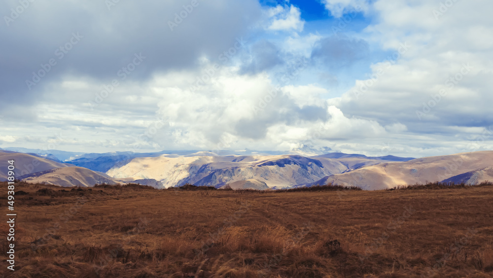 Brown autumn field in the highlands of the Caucasus overlooking a mountain valley and lush clouds in the blue sky