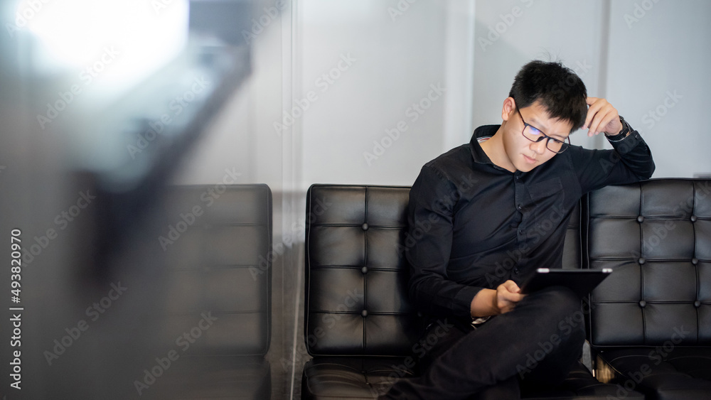 Young Asian businessman using digital tablet on leather chair in office. Male entrepreneur reading news on social media app. Online marketing and Big data technology for E-commerce business. 