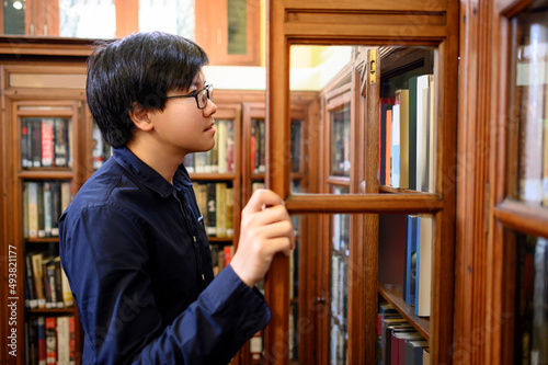Smart Asian man university student opening old wooden bookcase choosing vintage book in college library. Antique textbook resources for education research. History, Law and Literature learning concept