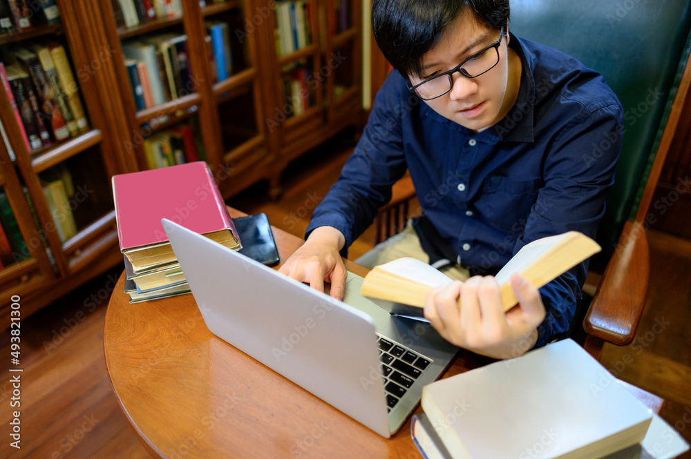 Asian man university reading book and using laptop computer nearby vintage bookcase or bookshelf in college library. Textbook resources for education research. Scholarship opportunity concept Asian man university reading book and using laptop computer nearby vintage bookcase or bookshelf in college library. Textbook resources for education research. Scholarship opportunity concept