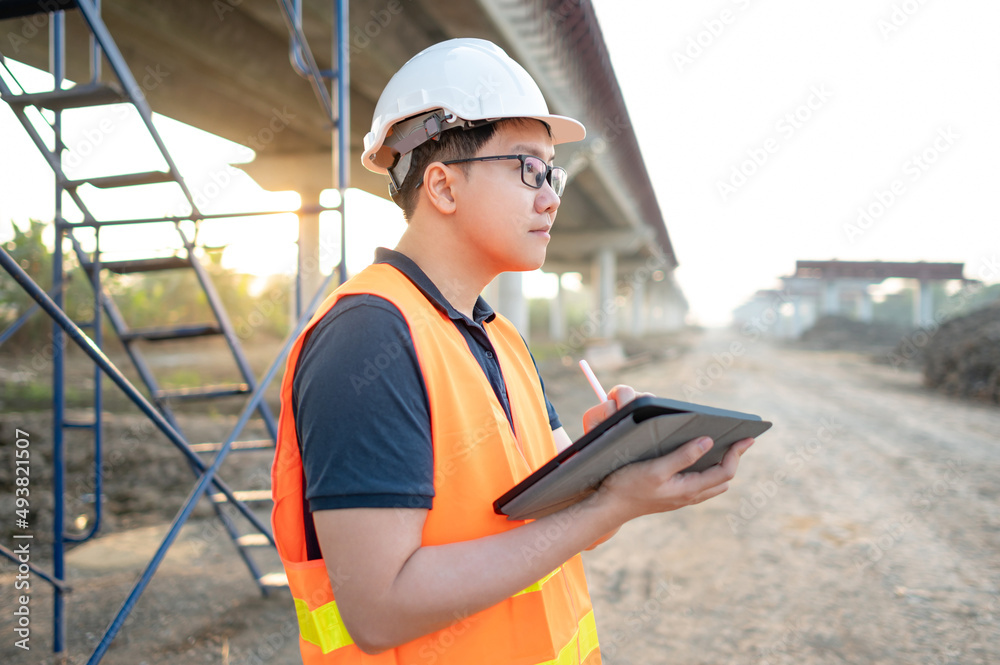 Smart Asian worker man or male civil engineer with protective safety helmet and reflective vest using digital tablet for project planning and checking architectural drawing at construction site. Smart Asian worker man or male civil engineer with protective safety helmet and reflective vest using digital tablet for project planning and checking architectural drawing at construction site.
