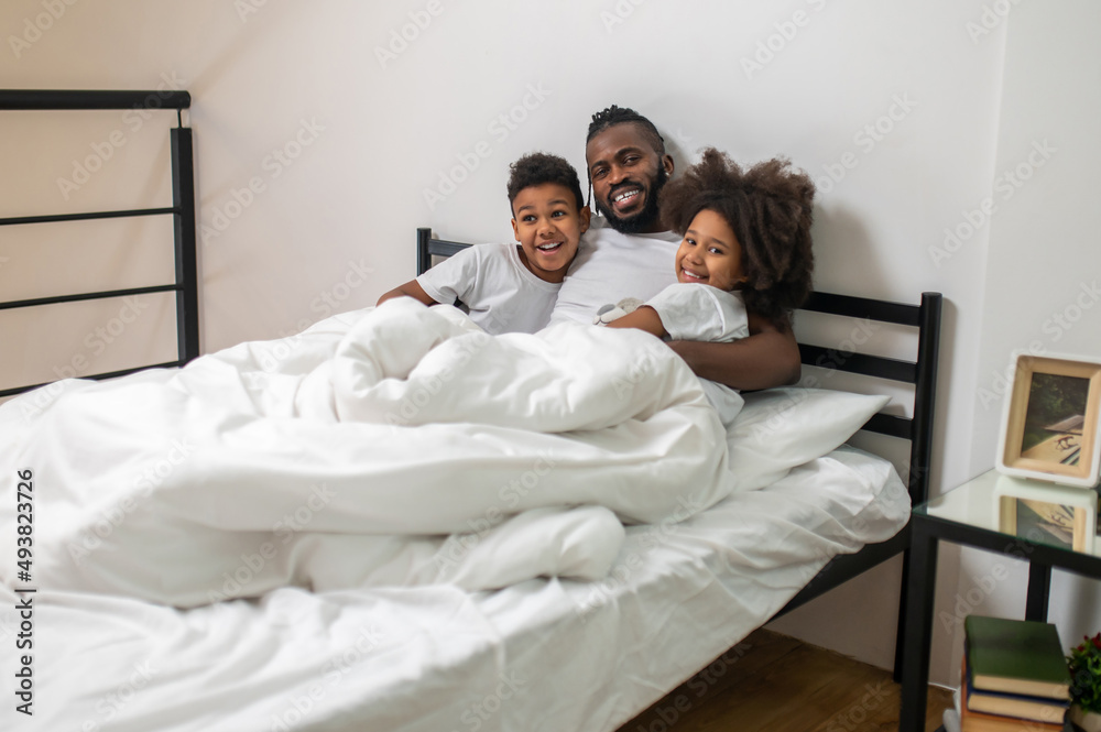 Man hugging kids in bed smiling at camera Man hugging kids in bed smiling at camera