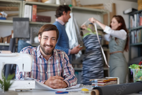 We can make anything. Portrait of a young fashion designer sitting in the workshop while his colleagues work in the background.