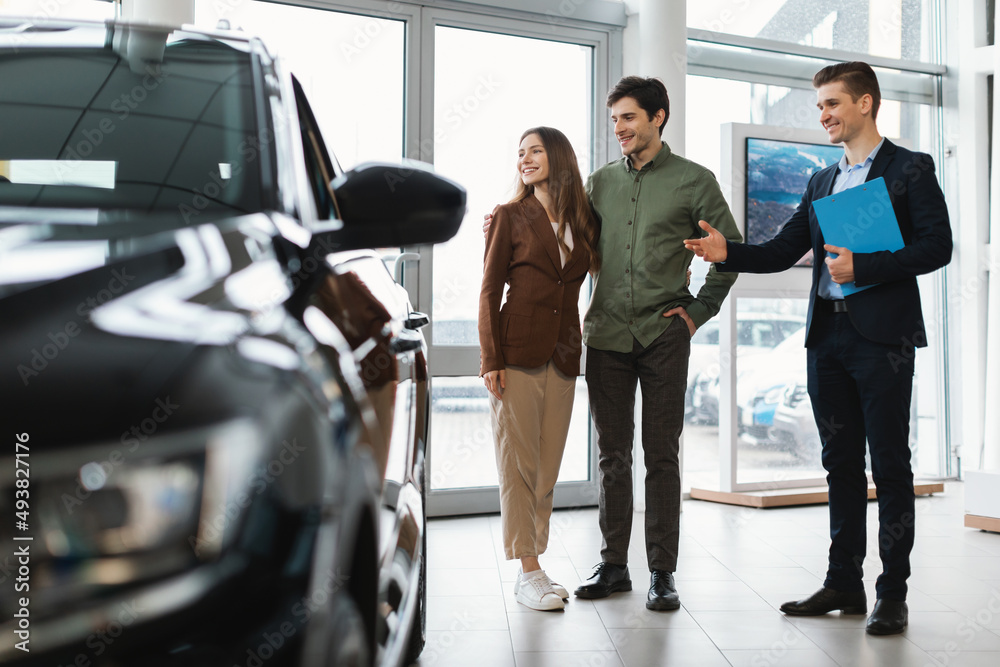 Happy car salesman showing new automobile to young couple at auto dealership