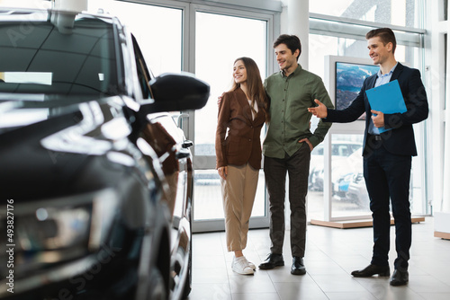Happy car salesman showing new automobile to young couple at auto dealership