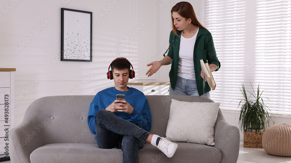 Obraz premium Strict mother with books scolding her son while he using smartphone at home. Teenager problems
