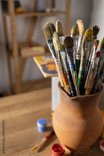 Artist supplies at wooden table. Paint brush in clay jug and art painter tools on wood background