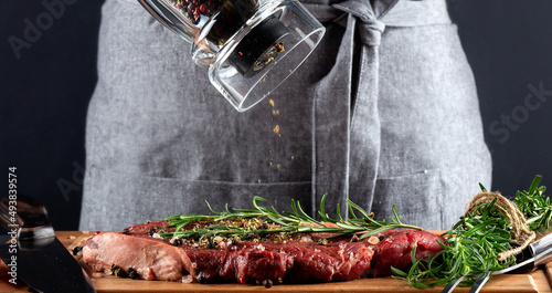 A woman cooks with peppers fresh raw ribeye steaks from marbled beef on a dark background. Nearby is a mixture of peppers, sea salt, garlic and rosemary. photo