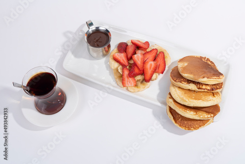 Cropped view of pancakes  banana and strawberry slices with hot chocolate and cup of tea on white plate shot on isolated area white background with selective focus from overhead angle.