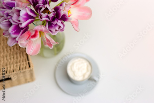 A bouquet of open pink and purple tulips, a wicker basket, a white cup with coffee on a white table.