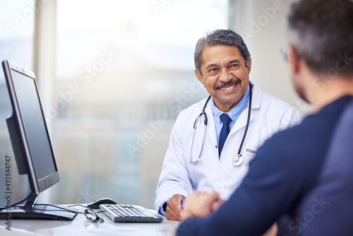 He gives all the right medical advice youll ever need. Shot of a cheerful mature male doctor seated at his desk while consulting a patient inside a hospital during the day.