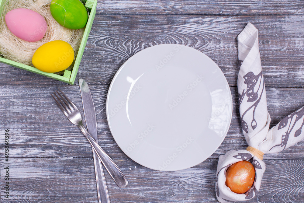 Φωτογραφία Easter composition with an empty white plate and an egg wrapped in a napkin in the form of rabbit ears, cutlery, colorful Easter eggs, spring leaves on a wooden gray table